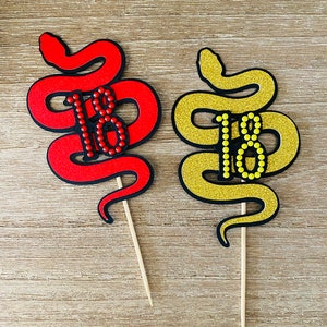 May include: Two cake toppers shaped like snakes with the number 18 in the centre. One snake is red and the other is gold glitter. Both snakes have red and yellow dots around the number 18.