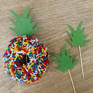 Puede incluir: Una dona de chocolate con chispas de colores coronada con un adorno de pastel de piña verde con purpurina. Se incluyen dos adornos de pastel de palmera verde con purpurina adicionales.