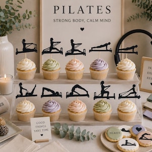 May include: A display of cupcakes with various pastel-colored frosting, each topped with a black silhouette of a Pilates exercise. The background includes a sign that reads "PILATES STRONG BODY, CALM MIND" and a small sign that reads "GOOD THINGS TAKE TIME".