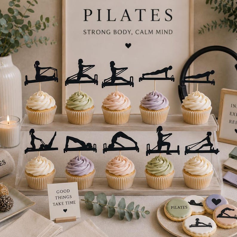 May include: A display of cupcakes with various pastel-colored frosting, each topped with a black silhouette of a Pilates exercise. The background includes a sign that reads "PILATES STRONG BODY, CALM MIND" and a small sign that reads "GOOD THINGS TAKE TIME".