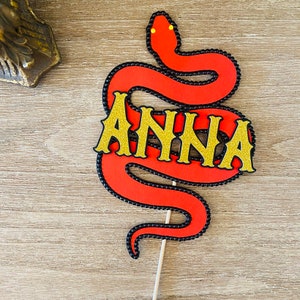 Puede incluir: Topper de pastel de serpiente roja con detalles de cuentas negras y el nombre "ANNA" en letras doradas con purpurina.