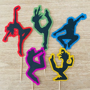Peut inclure: Cinq toppers de gâteau en silhouette de danseurs dans différentes poses. Les toppers sont en carton noir et sont de couleur rouge, vert, bleu, jaune et violet.