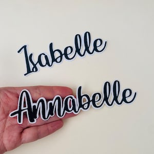 Puede incluir: Dos pegatinas de nombres en blanco y negro. Una pegatina dice "Isabelle" en una fuente cursiva. La otra pegatina dice "Annabelle" en una fuente cursiva. Las pegatinas tienen una fuente negra con un contorno blanco.