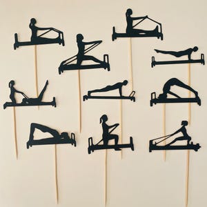 May include: A collection of black paper cutouts depicting various Pilates exercises on wooden skewers. The silhouettes show people using Pilates reformer machines in different poses, perfect for cake toppers or party decorations.