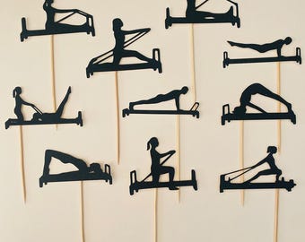 Puede incluir: Adornos para cupcakes con siluetas negras que representan varios ejercicios de Pilates en una máquina de reformer. Los adornos están sujetos a brochetas de madera y colocados sobre un fondo crema. Los adornos muestran diferentes poses.