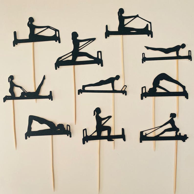 May include: A collection of black paper cutouts depicting various Pilates exercises on wooden skewers. The silhouettes show people using Pilates reformer machines in different poses, perfect for cake toppers or party decorations.