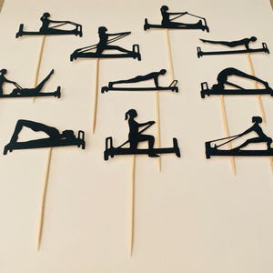 May include: Black paper cutouts of people doing Pilates exercises on wooden skewers. The cutouts depict various poses on Pilates equipment. The background is a light beige color. The cutouts are likely cake toppers or decorations for a fitness-themed event.