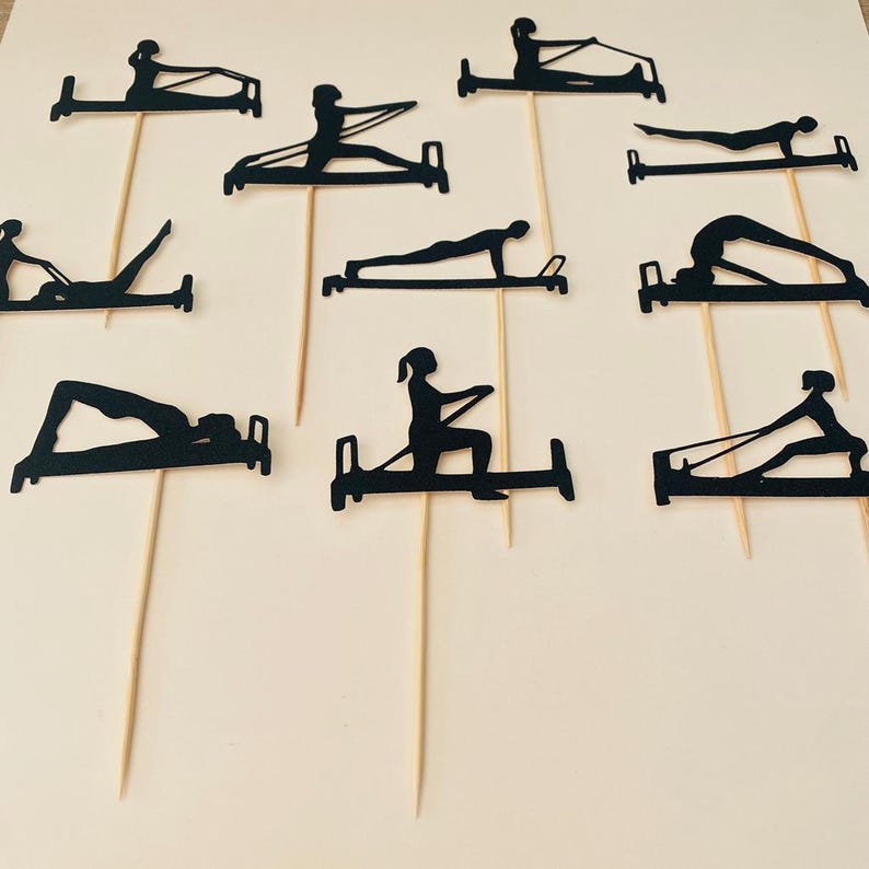 May include: Black paper cutouts of people doing Pilates exercises on wooden skewers. The cutouts depict various poses on Pilates equipment. The background is a light beige color. The cutouts are likely cake toppers or decorations for a fitness-themed event.