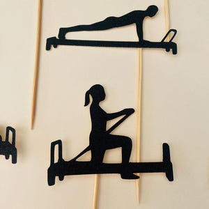 May include: Black silhouette cutouts of people exercising on Pilates equipment. The cutouts are attached to wooden skewers, suggesting they are cake toppers or decorative elements for a fitness-themed event.