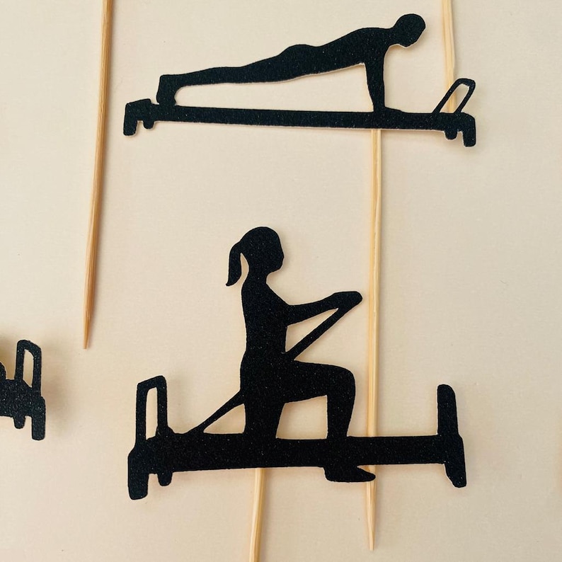 May include: Black silhouette cutouts of people exercising on Pilates equipment. The cutouts are attached to wooden skewers, suggesting they are cake toppers or decorative elements for a fitness-themed event.
