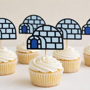May include: Cupcakes topped with white frosting and igloo-shaped toppers. The igloo toppers are light blue with black outlines and a dark blue door. The cupcakes have a light yellow cake base.