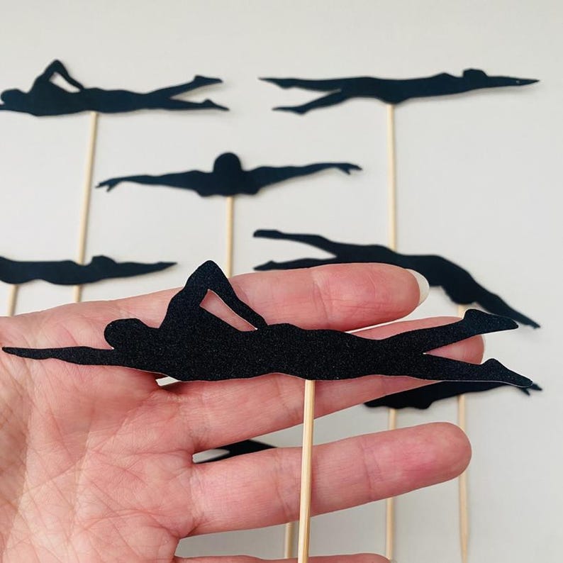 May include: Black silhouette cutouts of swimmers on wooden sticks. The swimmers are in various positions, with arms extended and legs kicking. These decorative toppers are ideal for a swimming-themed party or event.