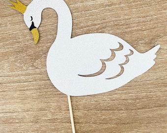 Puede incluir: Corte de papel blanco de un cisne con una corona dorada en la cabeza. El cisne tiene un ojo negro y un pico amarillo. El cisne está en un palo.