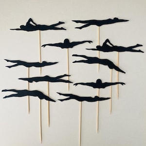 May include: A set of black swimmer silhouettes on wooden sticks. The swimmers are depicted in different strokes, with arms extended or streamlined. These toppers are suitable for a swimming-themed celebration or event.