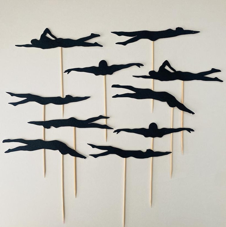 May include: A set of black swimmer silhouettes on wooden sticks. The swimmers are depicted in different strokes, with arms extended or streamlined. These toppers are suitable for a swimming-themed celebration or event.