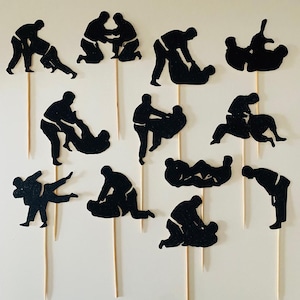 Puede incluir: Recortes de papel con purpurina negra de figuras de judo sobre palitos de madera. Las figuras representan varios movimientos y poses de judo. Estos adornos para tartas son ideales para una fiesta o evento temático de judo.