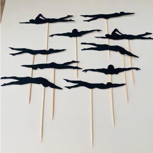 May include: A set of black swimmer silhouette cake toppers. Each topper depicts a swimmer in a streamlined pose, attached to a wooden skewer. The toppers are arranged on a white surface, ideal for a swimming-themed celebration.