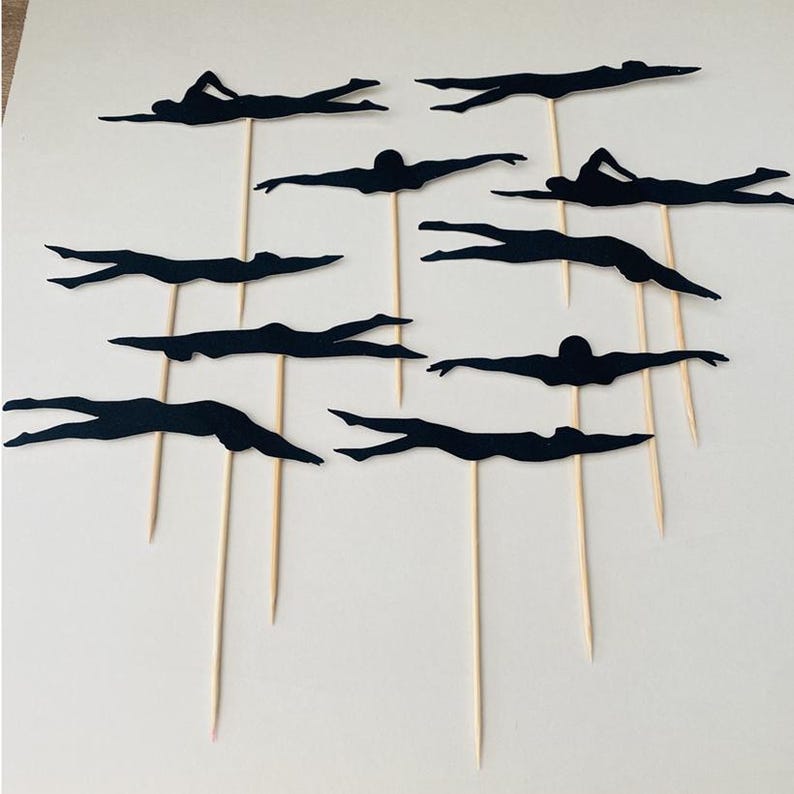May include: A set of black swimmer silhouette cake toppers. Each topper depicts a swimmer in a streamlined pose, attached to a wooden skewer. The toppers are arranged on a white surface, ideal for a swimming-themed celebration.