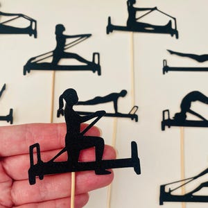 May include: Black silhouette cutouts of people doing Pilates exercises on wooden sticks. The cutouts depict various Pilates poses on the reformer machine. The background is a plain white surface. The cutouts are held by a hand.
