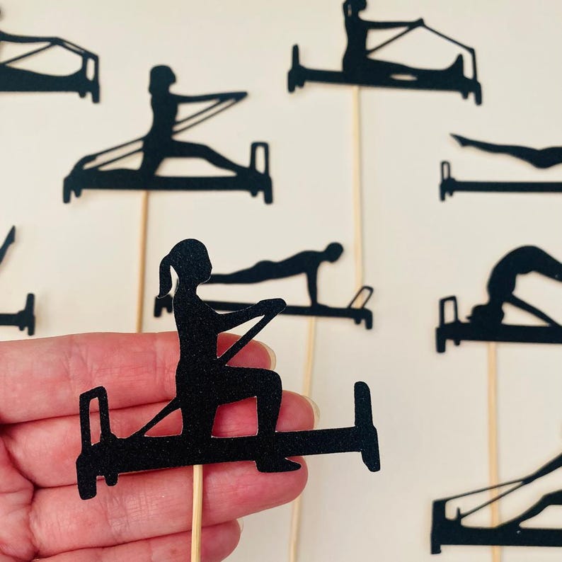 May include: Black silhouette cutouts of people doing Pilates exercises on wooden sticks. The cutouts depict various Pilates poses on the reformer machine. The background is a plain white surface. The cutouts are held by a hand.