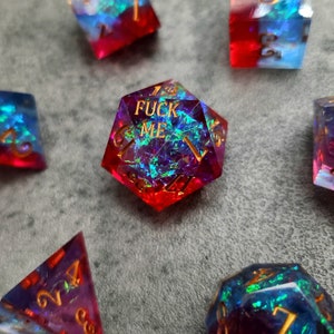 May include: A set of polyhedral dice featuring a translucent red and blue resin base with embedded iridescent foil flakes. The twenty-sided die prominently displays the text FUCK ME in gold lettering, matching the gold numbers on all other dice.