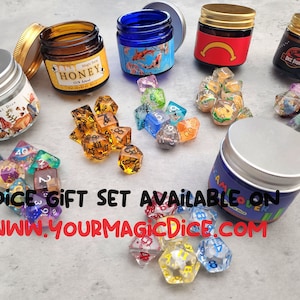 May include: Six colorful metal containers with lids, each containing a set of polyhedral dice. The containers have various designs, including a honey jar, a mushroom forest, a dinosaur, a red and yellow design, and a blue design with the text "Game Over". The dice are translucent and have different colors and numbers. The text "6 DICE GIFT SET AVAILABLE ON WWW.YOURMAGICDICE.COM" is printed on the table.