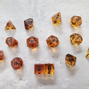 May include: A set of 11 amber dice with black numbers. The dice are in various shapes, including a d4, d6, d8, d10, d12, and d20.