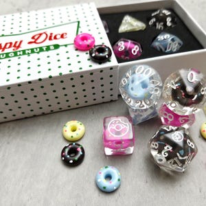 May include: A box of dice with a Krispy Kreme Doughnuts theme. The box is white with green polka dots and the words "Krispy Dice Doughnuts" in red and green. The dice are clear, pink, and black, with donut-shaped dice and miniature donuts.