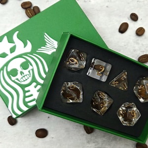 May include: A green box with a Starbucks-inspired logo featuring a skull, containing a set of seven clear dice with gold numbering. Coffee beans are scattered around the box.