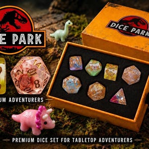 May include: A set of polyhedral dice featuring dinosaur-themed inclusions, housed in an orange box labeled Dice Park. The set includes a six-sided die with footprints, a twenty-sided die, and various other shapes, accompanied by small plastic dinosaur figurines.