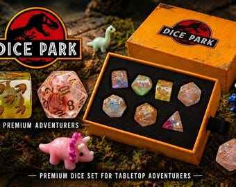 May include: A set of polyhedral dice in an orange box labeled DICE PARK, featuring dinosaur-themed inserts. Includes a pink triceratops, a blue dinosaur, a light green long-neck dinosaur, and various resin dice with gold flakes and fossil motifs.