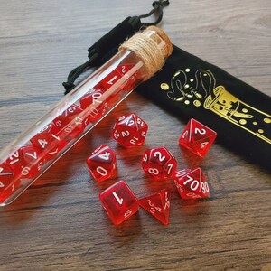 May include: A glass test tube filled with red dice with white numbers, a black velvet bag with a gold design of a test tube with bubbles, and nine red dice with white numbers on a wooden surface.