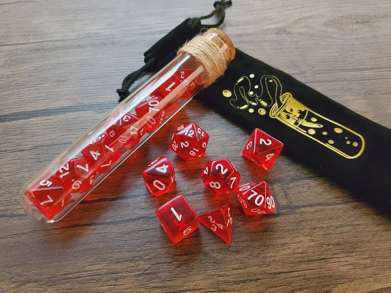 May include: A glass test tube filled with red dice with white numbers, a black velvet bag with a gold design of a test tube with bubbles, and nine red dice with white numbers on a wooden surface.