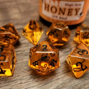 May include: A set of seven honey-colored dice with black numbers and a bee design on one die. The dice are on a wooden surface.