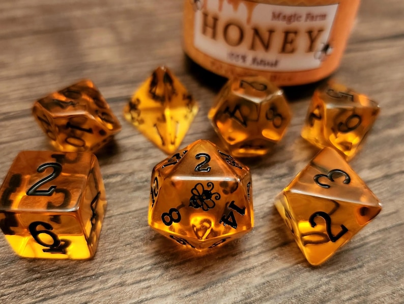 May include: A set of seven honey-colored dice with black numbers and a bee design on one die. The dice are on a wooden surface.
