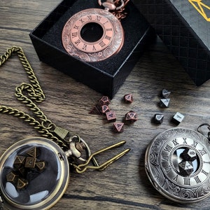 May include: Two antique-style pocket watches with chains, one with a copper finish and the other with a silver finish. Both watches have Roman numeral faces and are open to reveal dice inside. The watches are on a wooden surface with a black box and dice scattered around.