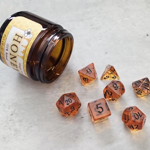 May include: A set of seven amber-colored dice with black numbers. The dice are scattered on a white surface. A small brown glass jar with a honey-themed label is tipped over on the left side of the image. The label reads "Honey 100% Real Polyhedral Dice".