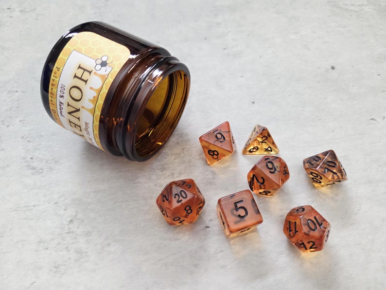 May include: A set of seven amber-colored dice with black numbers. The dice are scattered on a white surface. A small brown glass jar with a honey-themed label is tipped over on the left side of the image. The label reads "Honey 100% Real Polyhedral Dice".
