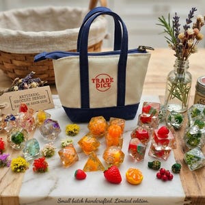 May include: A small canvas tote bag with navy blue handles and the red text TRADE DICE printed on the front. The bag is surrounded by various multi-sided resin dice containing embedded dried flowers, miniature fruit shapes, and greenery.