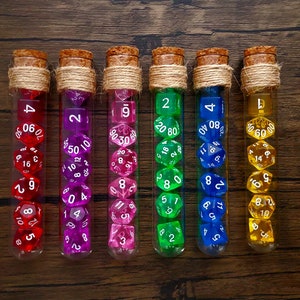 May include: Six glass test tubes filled with colorful dice. The dice are in various colors, including red, pink, purple, green, blue, and yellow. Each test tube is sealed with a cork stopper and tied with twine.