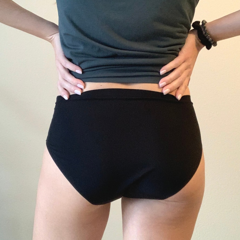May include: Black underwear with a high-waisted design. The underwear is made of a smooth, solid black fabric. The image shows the back of the underwear, with a person's hands on their hips. The background is a plain, light-colored wall.