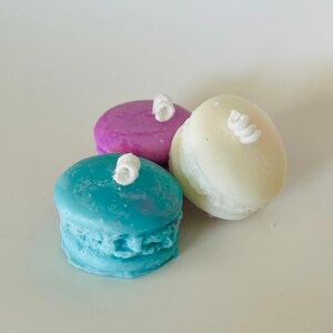 Könnte beinhalten: Drei bunte Macaron-förmige Kerzen mit weißem Frosting auf der Oberseite. Die Kerzen sind blau, lila und weiß.