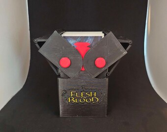 Peut inclure: Une boîte de jeu de cartes en impression 3D noire avec des boutons rouges et le texte "Flesh and Blood" en lettres dorées.