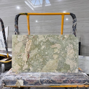 May include: A rectangular slab of green and beige marble with a mottled pattern, resting on a yellow and black wheeled cart. The marble has veins of red and white running through it. The cart has a floral patterned surface.