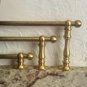 May include: Three horizontal brass rods of varying lengths, each mounted on a vertical support post with a circular base and a spherical finial at the end. The metal has a polished finish, and the rods are arranged in a stepped, tiered formation.