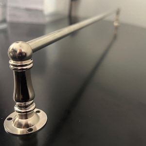 May include: A polished nickel towel bar with a decorative, rounded base and a long, straight bar. The bar is mounted on a dark surface, showcasing its shiny finish. The design features a classic, elegant style.