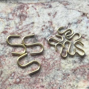 May include: An assortment of gold-colored metal hardware, including S-hooks and chain. The S-hooks are in various sizes and shapes, while the chain is a standard link design. The items are displayed on a speckled, light brown surface.