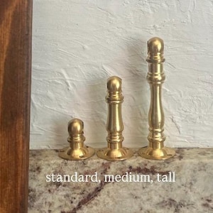 May include: Three brass door stops in varying heights, labelled standard, medium, and tall. Each piece features a cylindrical base with screw holes and a rounded top finial, showcasing a polished metallic gold finish and a smooth, turned design.