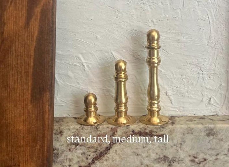 May include: Three brass door stops in varying heights, labelled standard, medium, and tall. Each piece features a cylindrical base with screw holes and a rounded top finial, showcasing a polished metallic gold finish and a smooth, turned design.