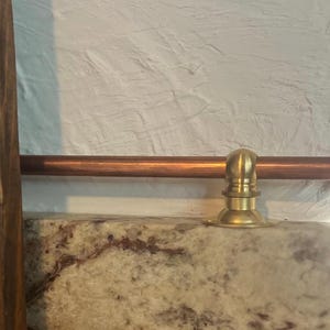 May include: A horizontal copper rod featuring a polished brass mounting bracket and a matching spherical end cap. The rod is positioned above a speckled granite surface, with a dark wooden vertical element visible on the left side of the frame.